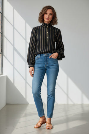 A woman with wavy brown hair, arms crossed, wears the MOMONI Thierry Blouse in lurex stripe gauze and a patterned skirt, standing against a pale background.
