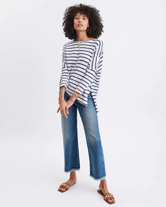 A woman with long, straight hair stands against a white wall, wearing the MERSEA Catalina Slub Tee in blue paired with white pants. The midweight textured cotton tee is from MERSEA. She looks at the camera with a neutral expression.