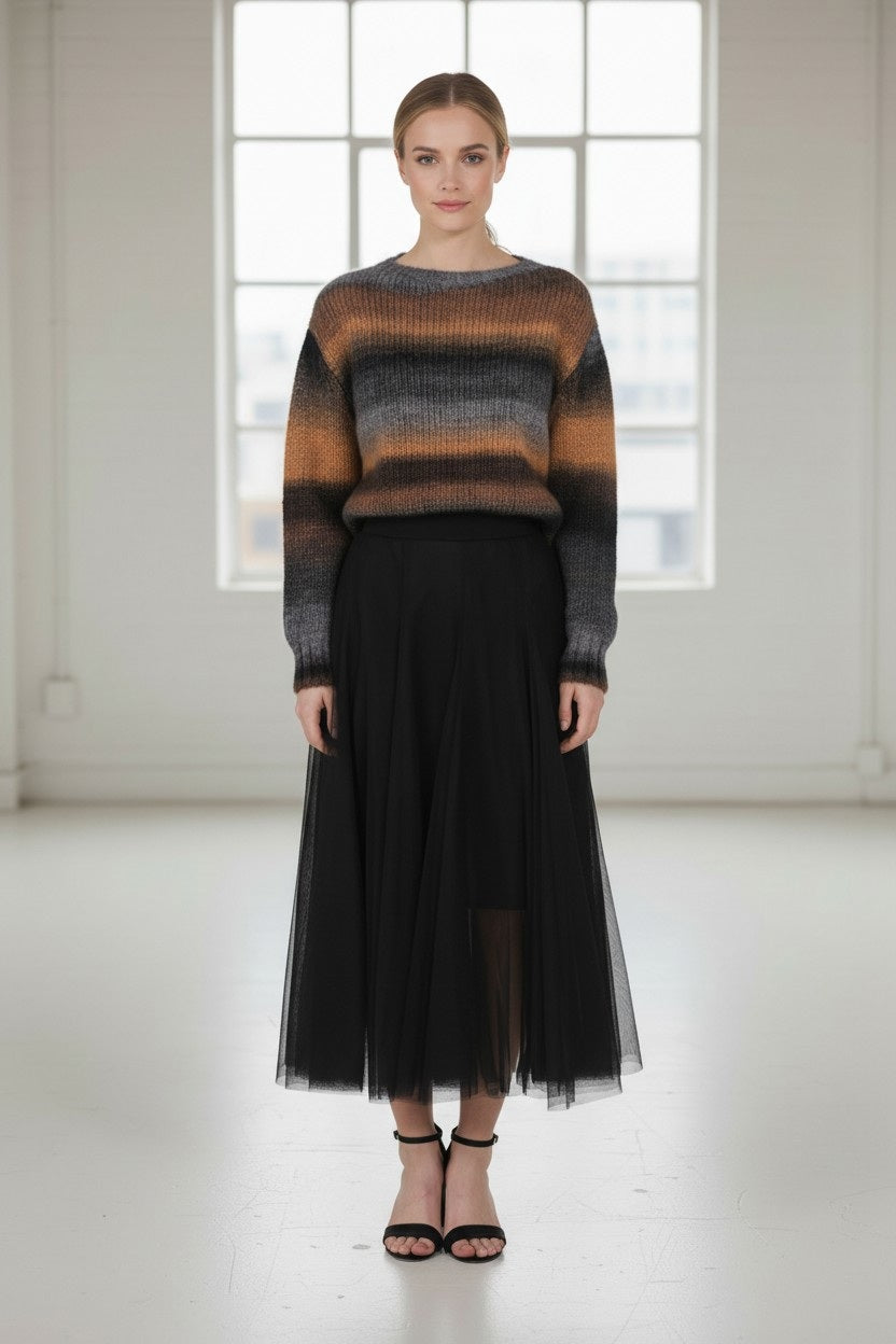 A woman stands indoors in front of a large window with soft natural light, wearing the JESSIE LIU Tulle Midi Skirt with Merino Wool Lining in Black, paired with a striped brown and gray sweater and black heels.