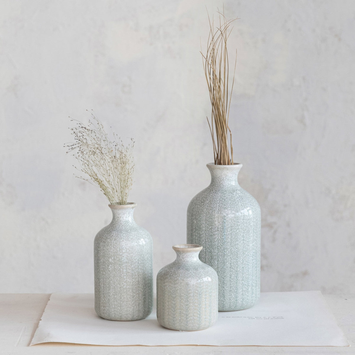 The CREATIVE COOP GRAY EMBOSSED STONEWARE VASE, MEDIUM features a tall, cylindrical shape with a narrow neck, flared rim, and textured leaf-like pattern—ideal as a decorative farmhouse accent.