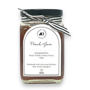 The FEARRINGTON SIGNATURE COLLECTION - FEARRINGTON HOUSEMADE PEACH JAM comes in a glass jar featuring a black lid and is adorned with a black and white striped ribbon. The white label reads "Peach Jam" and lists the ingredients as peach, vanilla, lemon, pectin, and sugar. Perfect as a hostess gift, it also notes that it is handmade by FEARRINGTON HOUSE and may contain allergens.