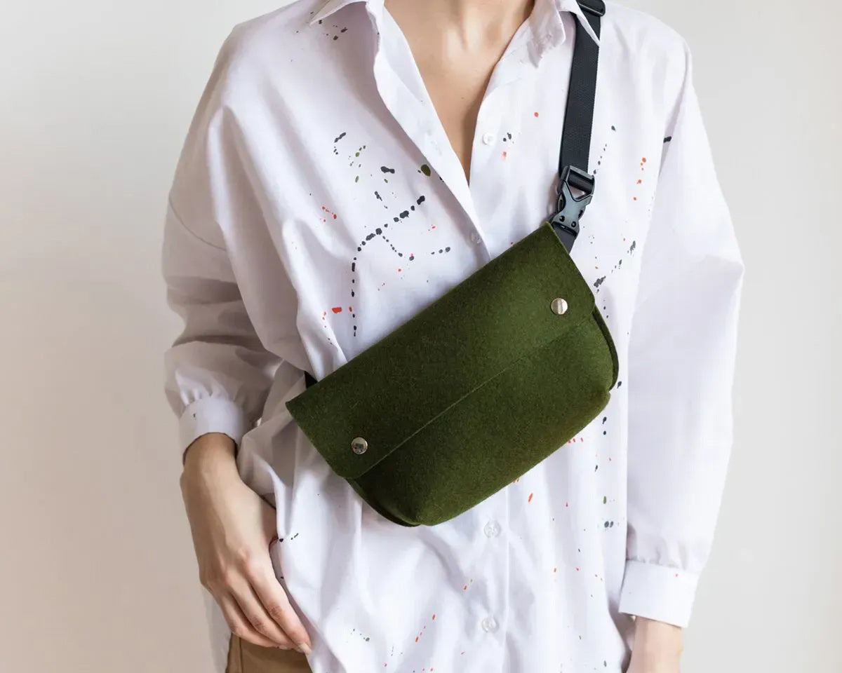 The WOOLBERRY - FELTED WOOL BELT BAG by WOOLBERRY is a small green belt bag crafted from felted wool, featuring a flap secured by two silver snap buttons, and is displayed against a white background.