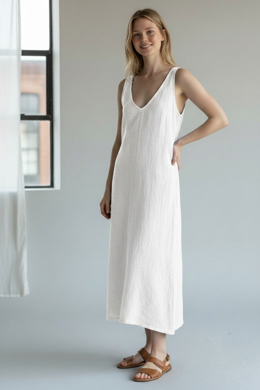 A woman stands indoors, smiling with one hand on her hip, wearing the LLEA BLUE - ERICA Linen Bias Cut Dress in White and brown sandals. She is positioned in front of a window adorned with sheer white curtains.