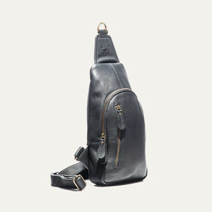 The WILL LEATHER GOODS - ZIP AROUND SLING BAG IN BLACK is a full-grain leather sling bag with gold zippers and buckle, small front pocket, adjustable strap, and top hanging loop, shown against a plain white background.
