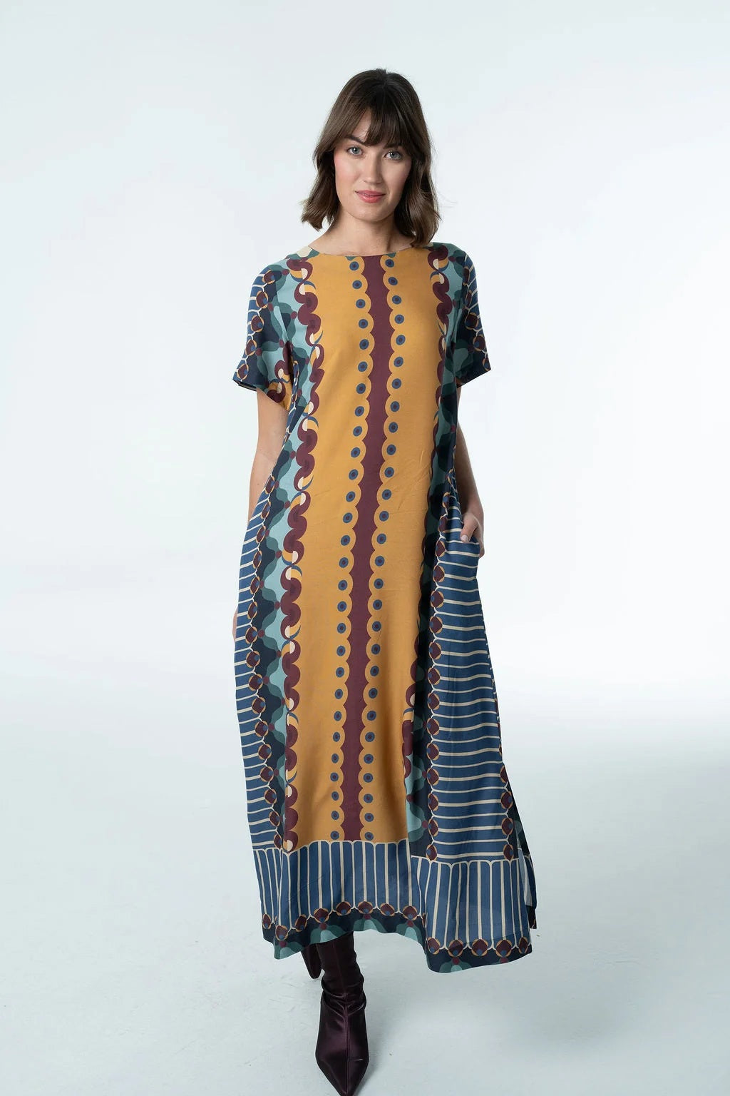 A woman wears the BRITON COURT - DIANA MAXI DRESS by BRITON COURT, a loose-fitting viscose maxi dress featuring vertical stripes in yellow, blue, and brown, short sleeves, practical side pockets, and an ankle-length hem. She is standing against a plain white background and pairs the dress with dark ankle boots.