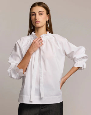 A woman with long brown hair models the HINSON WU ALINA LONG SLEEVE SHIRT, a bright blue blouse with a bow neck and ruffled sleeves, paired with dark pants. She stands against a light background, looking at the camera.