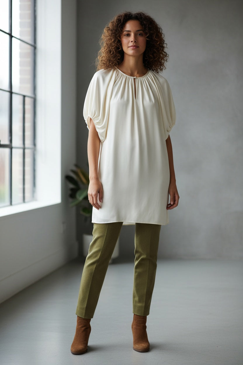 A woman stands indoors by a large window, dressed in the MOMONI - OLIVIA DRESS from MOMONI. The elegant white dress features gathered sleeves reminiscent of silk satin crepe. She pairs it with brown ankle boots, while the minimal background includes a gray wall and a partially visible plant.