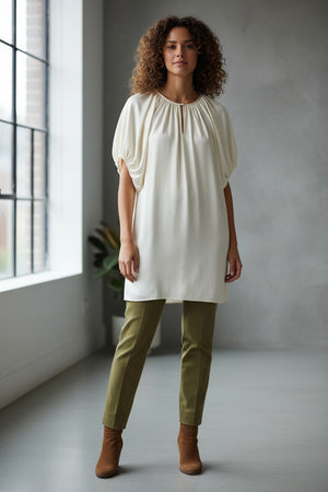 A woman stands indoors by a large window, dressed in the MOMONI - OLIVIA DRESS from MOMONI. The elegant white dress features gathered sleeves reminiscent of silk satin crepe. She pairs it with brown ankle boots, while the minimal background includes a gray wall and a partially visible plant.