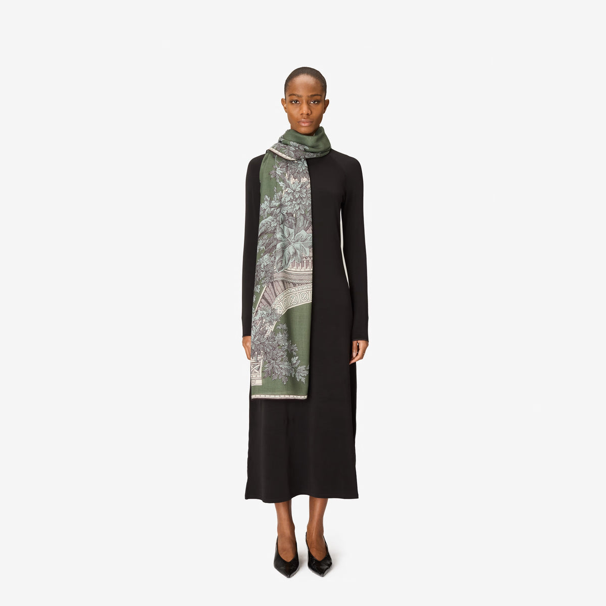 A person with short hair wears a long black dress and the INOUI EDITIONS 1889 WOOL SCAR FIN KHAKI scarf by INOUI—a green patterned accessory featuring intricate floral and geometric designs, crafted from 100% wool in India—while standing against a plain white background.