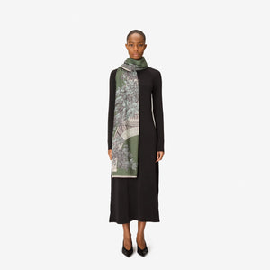 A person with short hair wears a long black dress and the INOUI EDITIONS 1889 WOOL SCAR FIN KHAKI scarf by INOUI—a green patterned accessory featuring intricate floral and geometric designs, crafted from 100% wool in India—while standing against a plain white background.