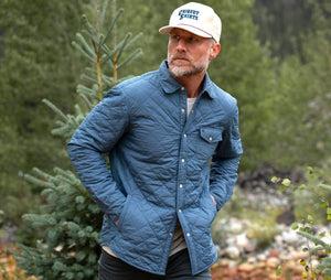 A man with a gray beard wears the CRIQUET - QUILTED SHACKET, a light blue piece with a pearl snap closure, along with a cream cap. He stands outdoors among pine trees and greenery in a forested area.