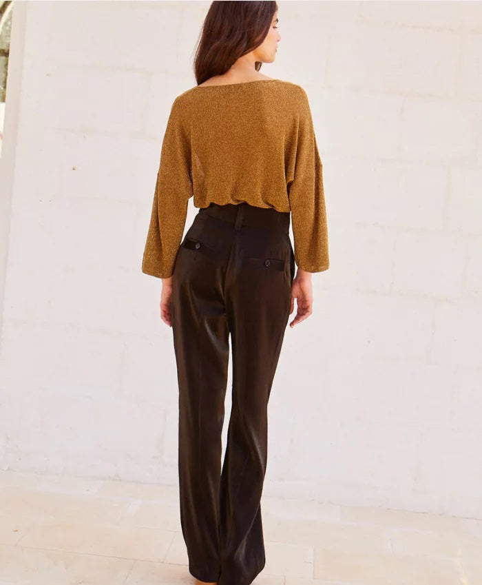 A person stands against a light-colored stone wall, wearing a loose-fitting, long-sleeved brown top with bio-galalite buttons paired with the high-waisted, wide-leg MANHATTAN PANT from MOMONI. They have long hair and are looking directly at the camera. The background is simple and uncluttered.