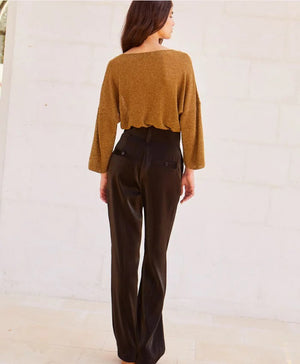 A person stands against a light-colored stone wall, wearing a loose-fitting, long-sleeved brown top with bio-galalite buttons paired with the high-waisted, wide-leg MANHATTAN PANT from MOMONI. They have long hair and are looking directly at the camera. The background is simple and uncluttered.