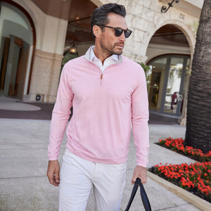 A man with short dark hair and a beard wears the COLLARS AND CO PALM PIMA BLEND QUARTER ZIP IN PINK, holding sunglasses in one hand. He stands outdoors by a building with arches, palm trees, and flowerbeds.