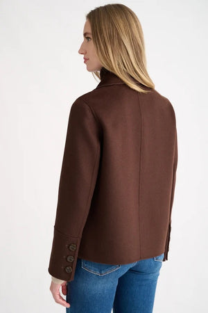 A close-up shows a person wearing the KINROSS Knit Insert Notch Collar Coat in Chocolate, featuring a wide lapel and crafted from luxurious wool cashmere, layered over a dark brown high-neck zip-up sweater. Only the neck, shoulder, and part of the face are visible.
