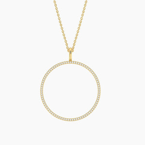 A woman wearing a cream square-neck top and the ELLA STEIN - YOU ARE MY EVERYTHING PENDANT NECKLACE by ELLA STEIN, featuring a large, circular, diamond-studded pendant. The natural diamond necklace adds an elegant touch to her look, complemented by her neatly tied-back hair and the neutral background.