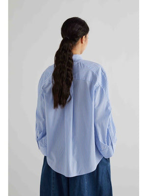 A woman with dark hair tied back models the MEIMEIJ STRIPE COTTON SHIRT—an oversized light blue and white striped cotton top with wide sleeves—paired with blue pants, standing against a plain light gray background.