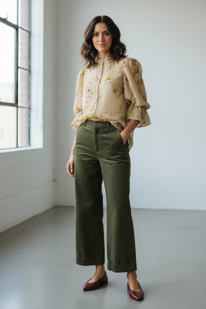 A woman stands in a sunlit room, smiling with her hands relaxed by her sides, wearing the MIRTO 1956 - BLOUSE WITH FLORAL PRINT from MIRTO 1956, paired with blue jeans and brown sandals.