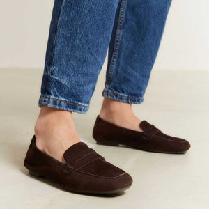 A person stands on a light floor wearing rolled-up blue jeans and REQINS HEMA suede loafers in Marron, showcasing the artisanal craftsmanship of REQINS.