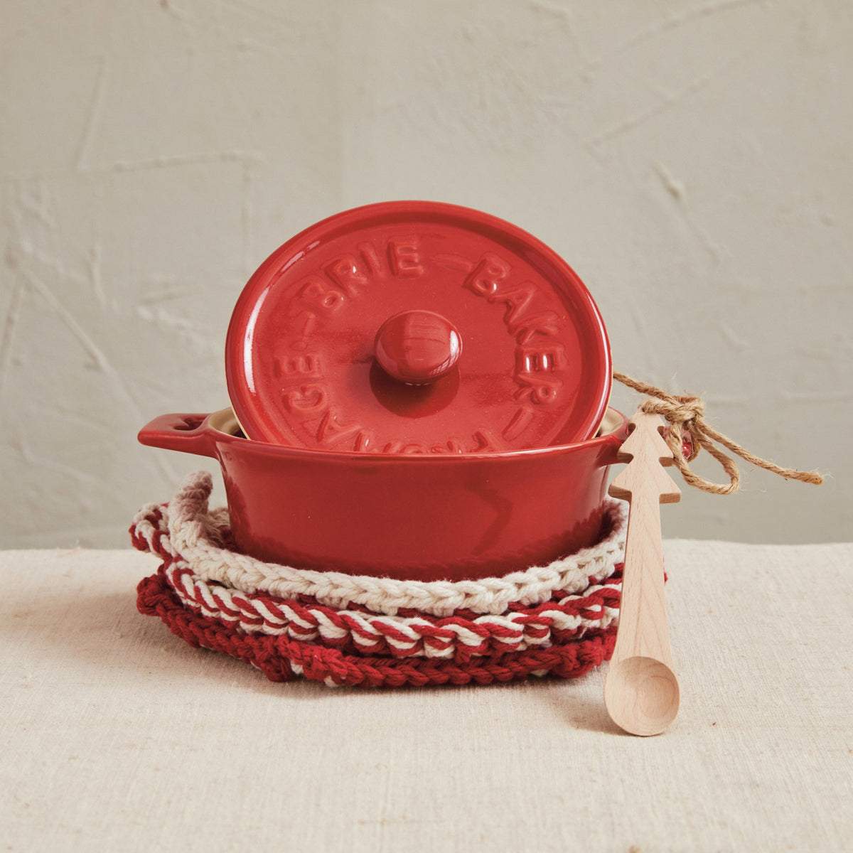 The CREATIVE COOP STONEWARE BRIE BAKER WITH BAMBOO TREE SPOON sits atop red and white pot holders, its lid angled nearby, with a spoon tied in twine on a beige fabric surface against a textured light wall.