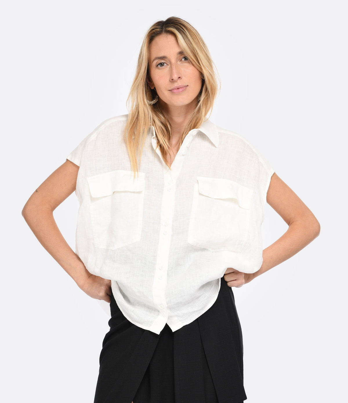 A woman with long blonde hair models the MOMONI SLEEVELESS LINEN SHIRT, a loose-fitting, garment-dyed white button-up with two front pockets, paired with black pants, standing against a plain light background.