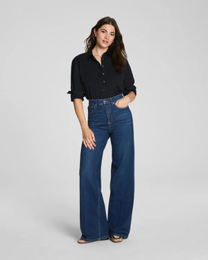 A person models the SPANX - SPANXSHAPE AUTHENTIC 360 WIDE LEG JEAN by SPANX, featuring a high-waisted fit and crafted from 4-way stretch denim, paired with a black shirt. Only the lower half of their body is visible as they stand with one hand at their side against a plain background.