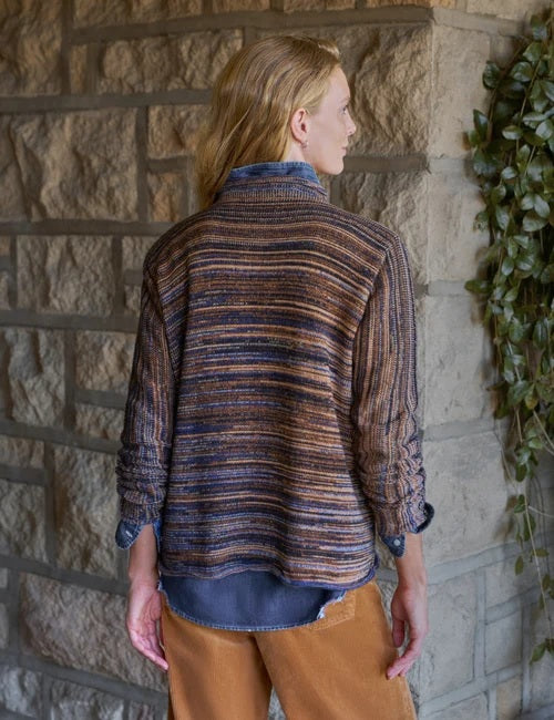 A woman stands near a window in a bright, minimal room, smiling and looking to the side while wearing the FRANK & EILEEN MONTEREY Rolled Funnel Neck Sweater in Pure Italian Cotton Harvest Stripes layered over a chambray shirt, paired with tan pants and brown ankle boots.