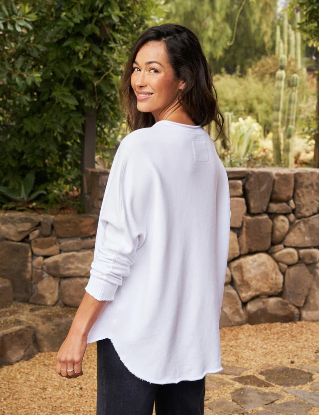 A person with long, wavy blonde hair is standing outdoors near a large tree, wearing the FRANK &amp; EILEEN ANNA LONG-SLEEVE CAPELET in navy over a white shirt and blue pants. The background showcases green foliage and a stone path. The person is smiling and gazing to the side.