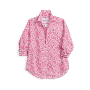 A woman stands on a brick path in a lush garden, wearing the FRANK & EILEEN EILEEN Relaxed Button Up in Liberty Print Pink Flowers, with blue jeans and woven flats. She carries a tote of pink flowers, surrounded by greenery and blooms.