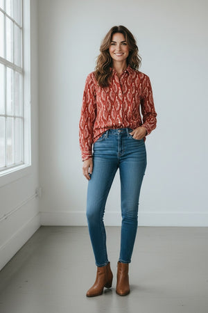 A woman with wavy brown hair smiles as she poses with one hand in her pocket, wearing the Hartford Cinema Shirt in Red Peacock Print—a loose-fitting, 100% cotton shirt featuring a vibrant red hue with a white peacock pattern—paired stylishly with blue jeans.