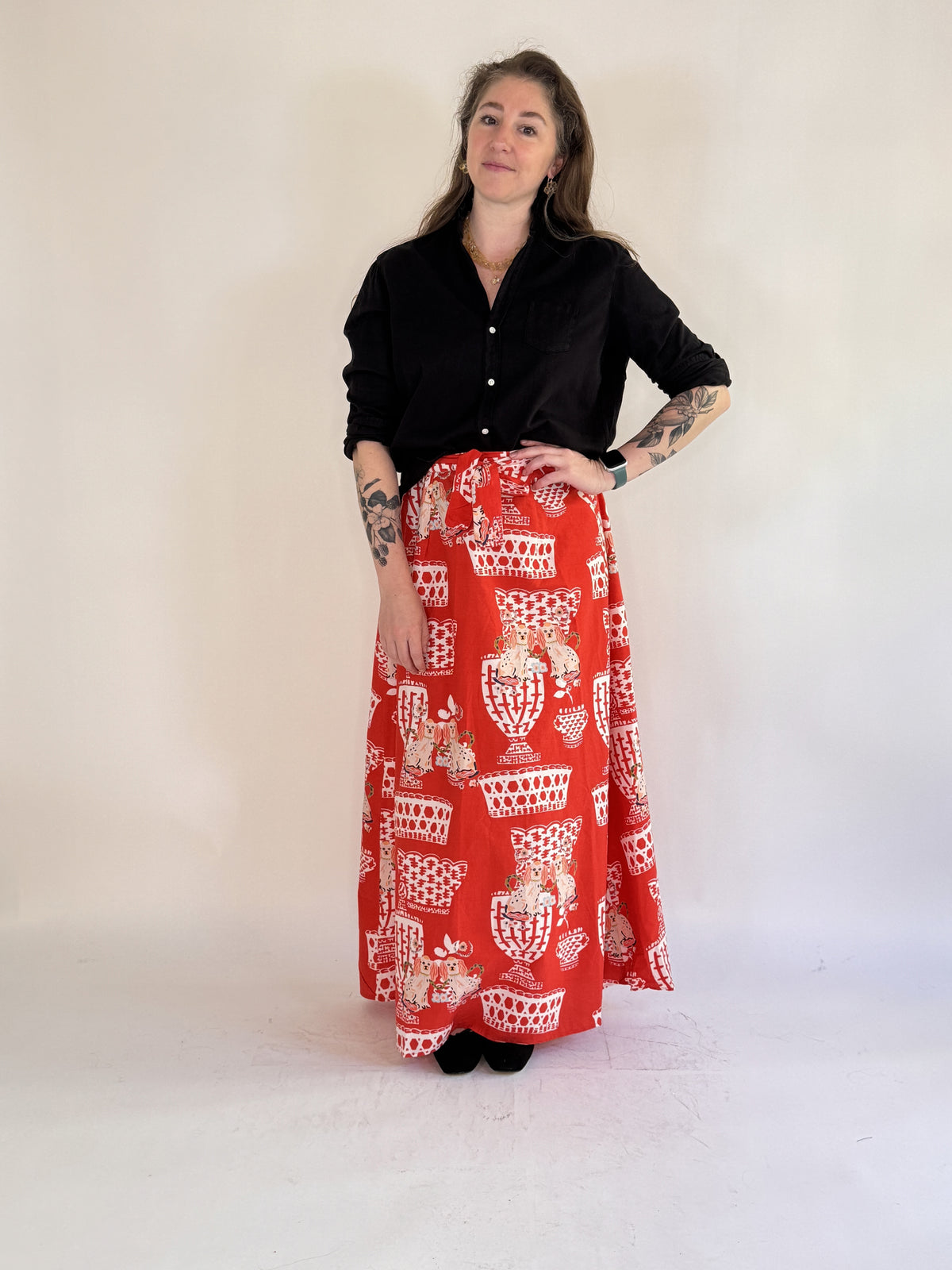 A person with tattooed arms stands against a light background, wearing a black top with sheer ruffled sleeves, black shoes, and the FARYLINA - FIFI WRAP SKIRT by FARYLINA featuring a white and beige cat pattern.