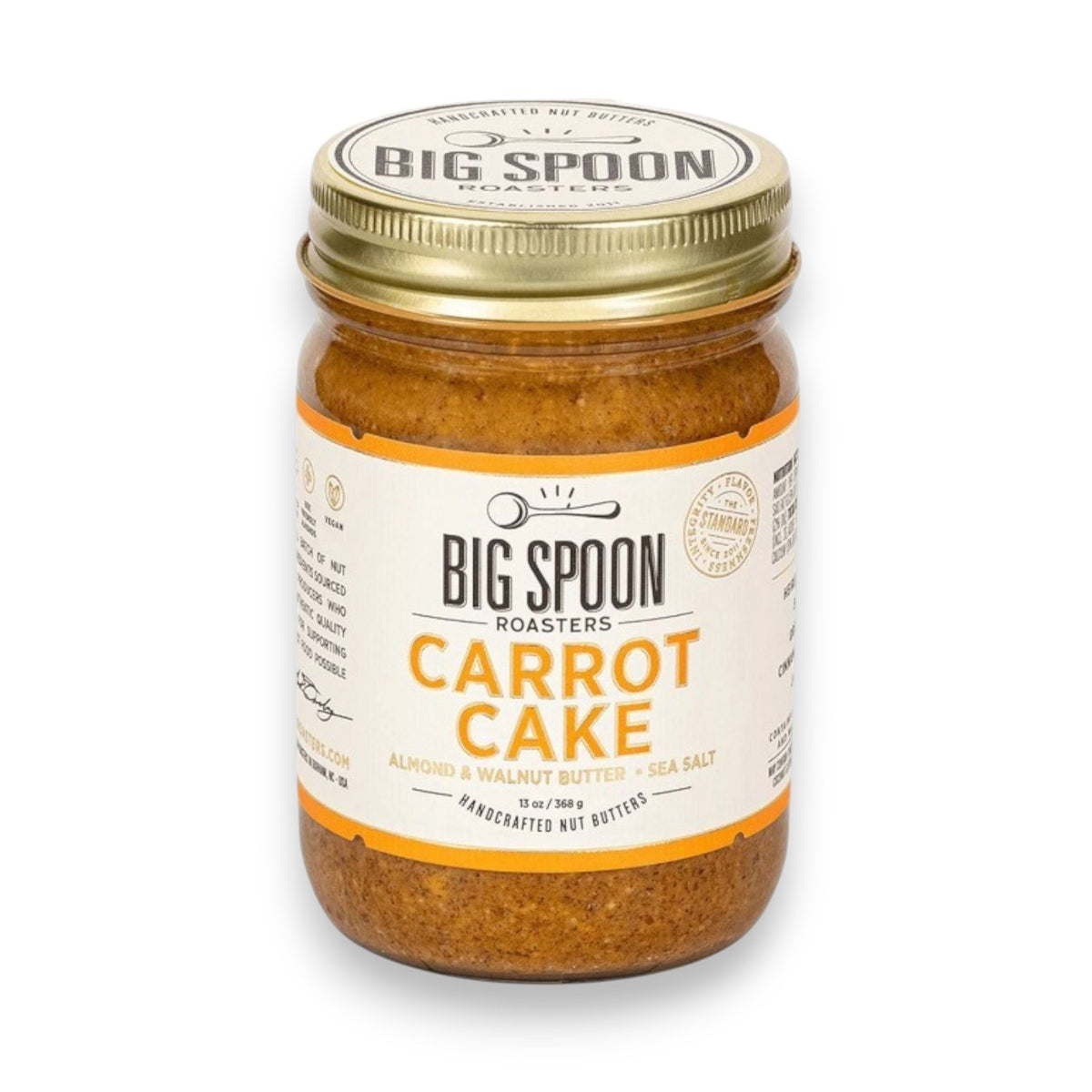 A jar of BIG SPOON ROASTERS - CARROT CAKE 13OZ, a vegan almond and walnut butter with no refined sugar, sits with a spoon inside in front of a frosted carrot cake and a plate holding a slice of the cake.