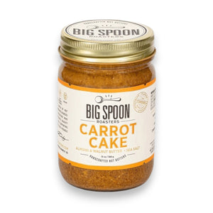 A jar of BIG SPOON ROASTERS - CARROT CAKE 13OZ, a vegan almond and walnut butter with no refined sugar, sits with a spoon inside in front of a frosted carrot cake and a plate holding a slice of the cake.