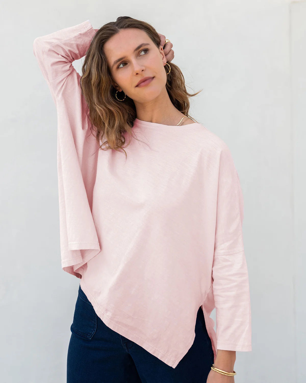 A woman with wavy brown hair models the MERSEA - CATALINA SLUB TEE—an oversized, long-sleeved white top with thin gray stripes—by MERSEA. She pairs it with blue jeans and stands against a plain, light background, hands behind her back.