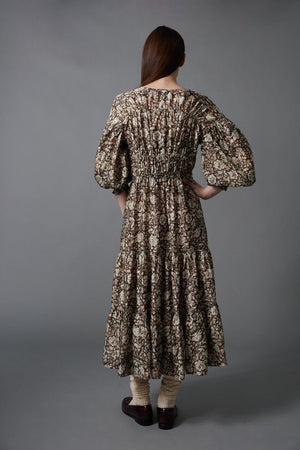 A woman stands against a gray background wearing the HOPE FOR FLOWERS TIERED PEASANT DRESS with puffed sleeves and a shirred bodice. She has her hands in its pockets and pairs dark loafers with light-colored socks.