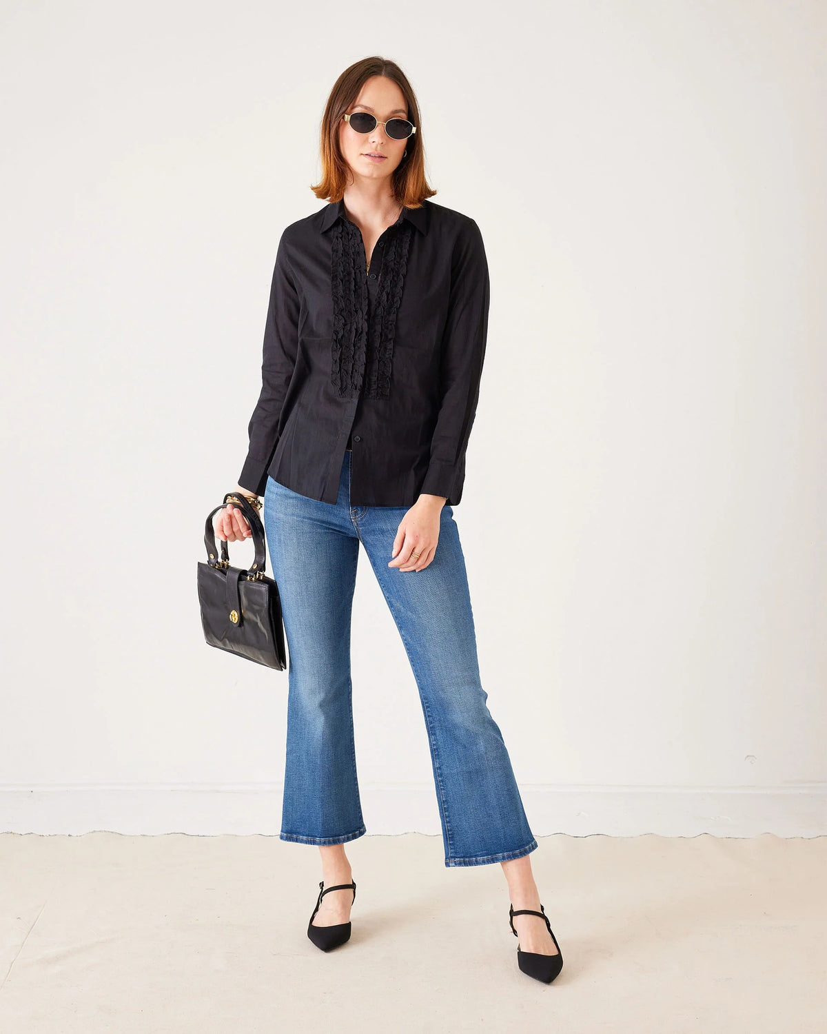 A woman stands against a plain white background wearing sunglasses, a black long-sleeve blouse, black heels, and holding a black handbag. She completes her look with the MERSEA Murphy Mini Boot Jean in Tide from MERSEA—blue, cropped organic cotton jeans designed for a gap-free waist.
