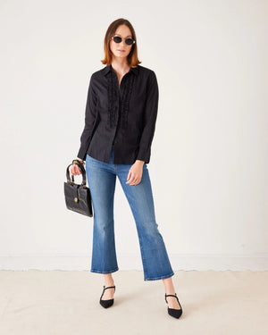A woman stands against a plain white background wearing sunglasses, a black long-sleeve blouse, black heels, and holding a black handbag. She completes her look with the MERSEA Murphy Mini Boot Jean in Tide from MERSEA—blue, cropped organic cotton jeans designed for a gap-free waist.