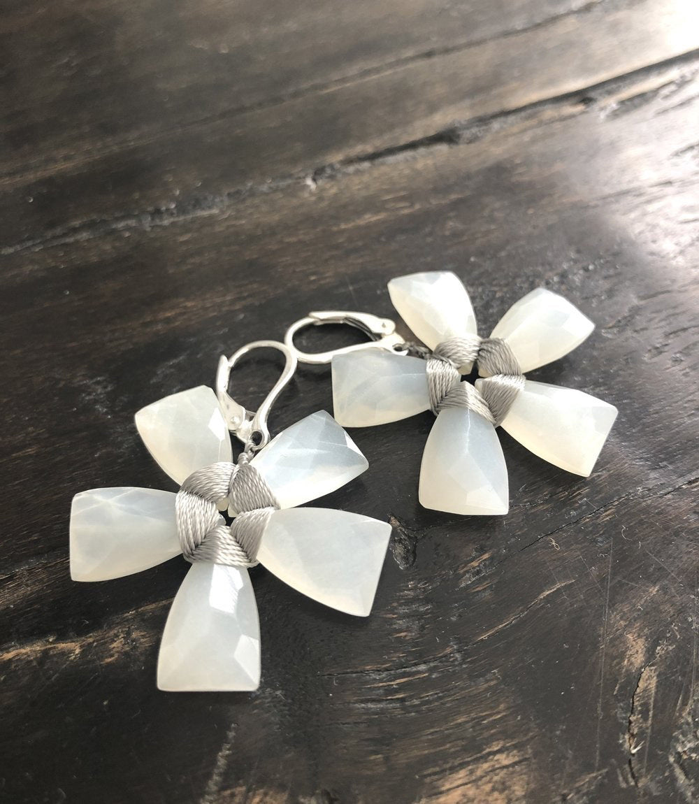 The ANN LIGHTFOOT - WHITE MOONSTONE FLOWER EARRINGS by ANN LIGHTFOOT are displayed on a dark wooden surface, showcasing their exquisite design with moonstones forming petal-like shapes and accented with metallic wire at the center.