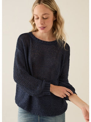 A woman with long blonde hair wears the BASE MILANO - CREWNECK COTTON BLEND SWEATER in navy, paired with light denim jeans. She stands against a plain background, looking down with a slight smile as she adjusts her sleeve.
