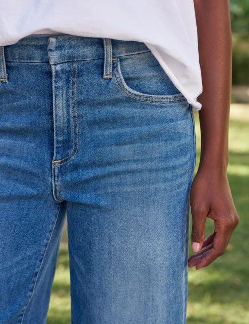 Standing outdoors on stone steps, a person wears the FRANK & EILEEN Galway Wide Leg Jean in 2002 Mid Wash—crafted from Italian Dream Denim with a hand-distressed raw hem—paired with a white shirt and tan sandals. Green grass and stone landscaping form the backdrop.