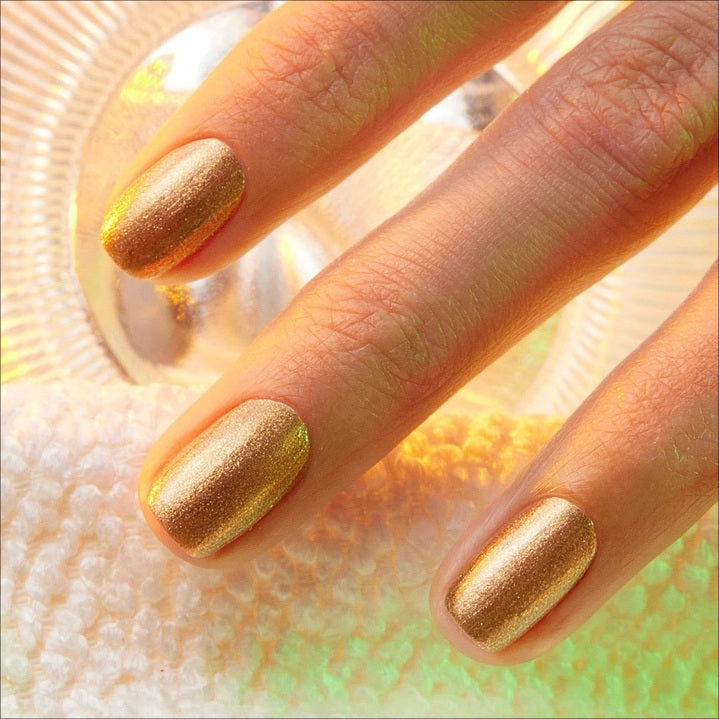 A hand adorned with metallic gold vegan nail polish holds a bottle of MANUCURIST - GREEN™ NAIL POLISH IN SHIMMER by MANUCURIST INC, set against a textured, light-filled background.