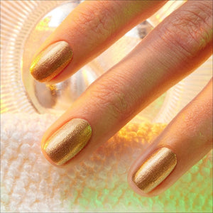 A hand adorned with metallic gold vegan nail polish holds a bottle of MANUCURIST - GREEN™ NAIL POLISH IN SHIMMER by MANUCURIST INC, set against a textured, light-filled background.