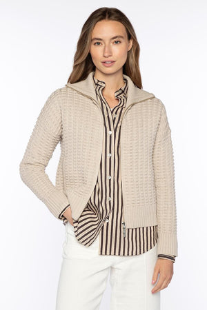 A woman wears the KINROSS Bobble Zip Cotton Cardigan in white over a black and beige striped button-up shirt with white pants, standing facing forward with one hand in her pocket against a plain white background.