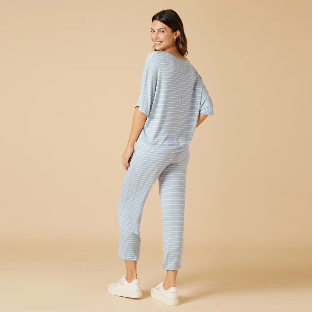 A woman wears the SOFTIES Dream Relaxed V-Neck with Capri Lounge Set in Sky Blue Stripe, featuring soft jersey fabric, short sleeves, matching drawstring capri pants, and white sneakers for a relaxed, all-day comfortable look.