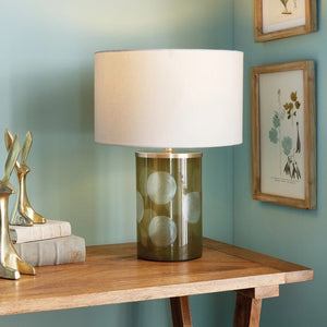 The NAPA HOME AND GARDEN - NORMAN LAMP, with a whimsical white drum shade and green polka-dotted cylindrical base, sits on a wooden table against a light blue wall decorated with botanical prints, surrounded by books and abstract sculptures.