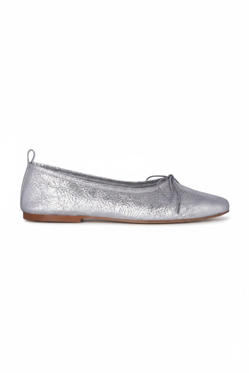 The AUGUSTA - SIENNA BALLET FLAT by AUGUSTA, handcrafted in Spain, showcases a silver finish with a front bow, low brown sole, and heel pull tab, displayed in side profile against a plain light background.