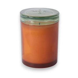 The MERSEA - BEACH BUNGALOW JOLIE JAR CANDLE by MERSEA is a burnt orange soy wax candle in a glass jar with a clear lid, photographed at an angle against a white background, evoking hints of eucalyptus and moss.