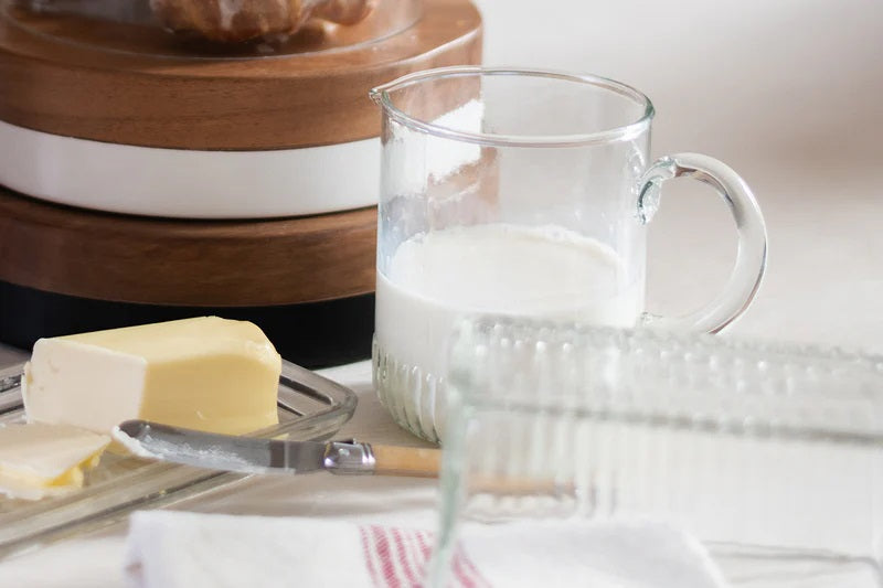 The ETUHOME - BISTRO GLASS MINI JUG by ETUHOME, filled with milk, rests on a white surface next to a butter dish and knife, with a clear glass baking dish and wooden container in the background.