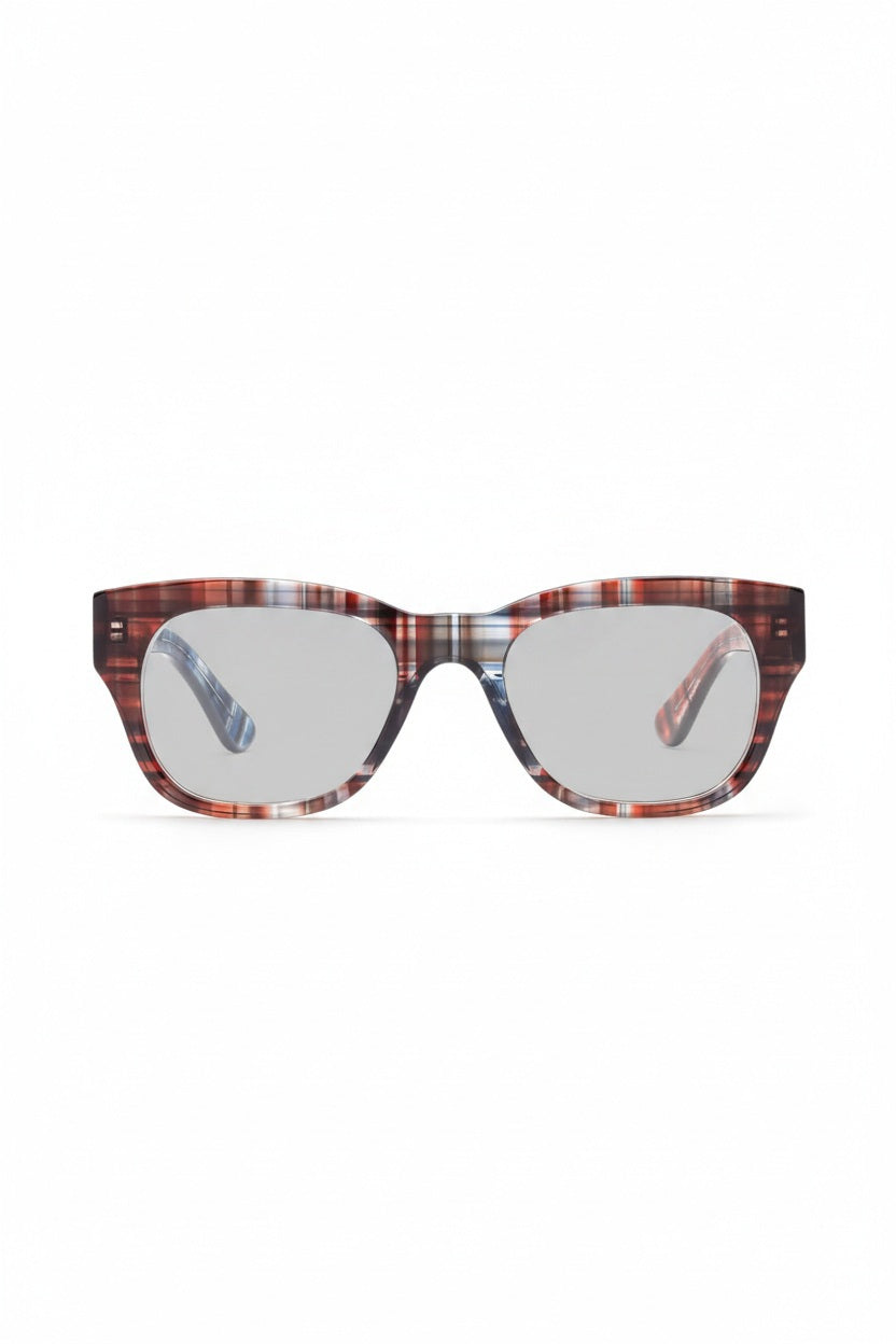 A person wearing CADDIS - MIKLOS READERS IN MADRAS PATCHWORK glasses with thick-rimmed, multicolored patterned frames against a solid red background.