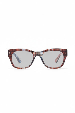 A person wearing CADDIS - MIKLOS READERS IN MADRAS PATCHWORK glasses with thick-rimmed, multicolored patterned frames against a solid red background.